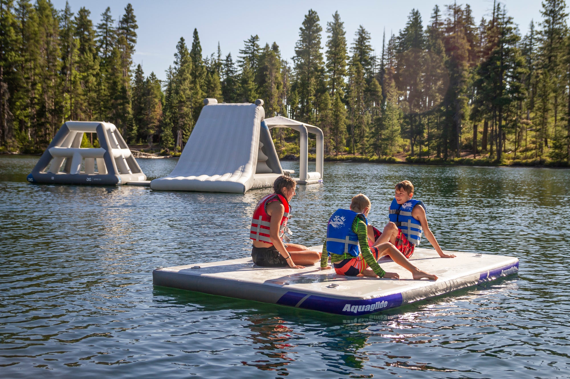 Three people in life vests sit on a floating mat on a lake with inflatable structures nearby.
