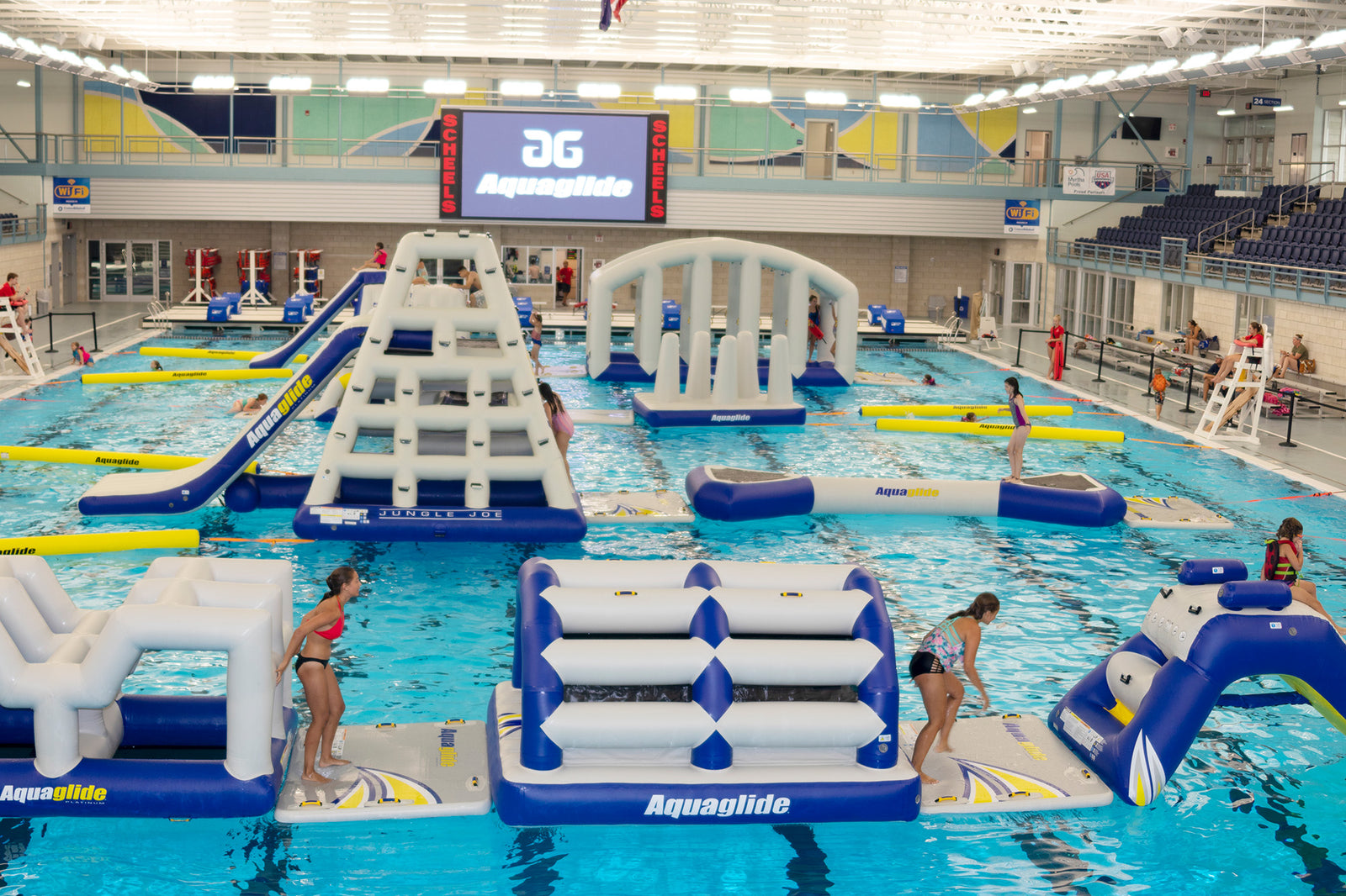 Indoor pool with inflatable obstacle course and people in swimwear.