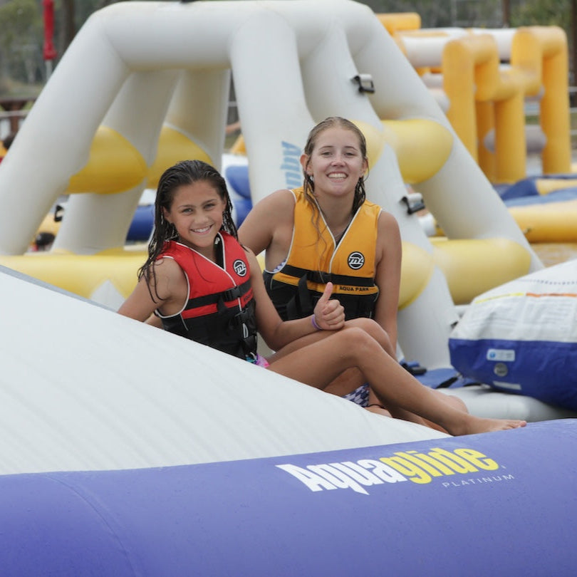 Two girls in life vests sit smiling on the Kaos inflatable with water park features behind them.