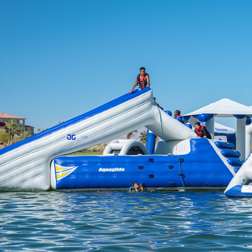 Kids in life vests climb and play on the Loki Corner inflatable slide over calm water.