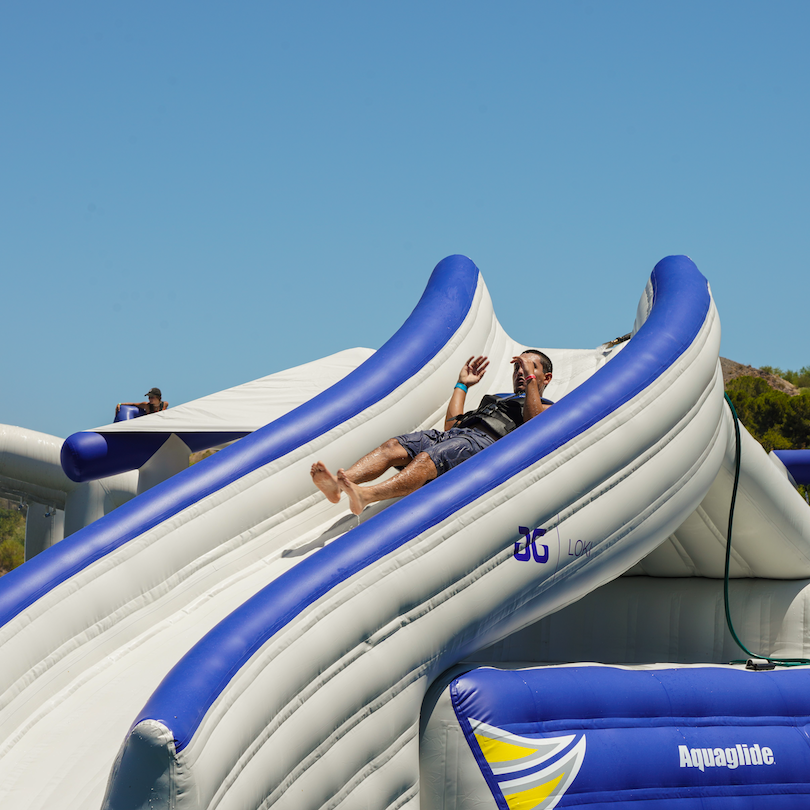 Man in a life vest slides down the Loki Corner inflatable under a clear blue sky.