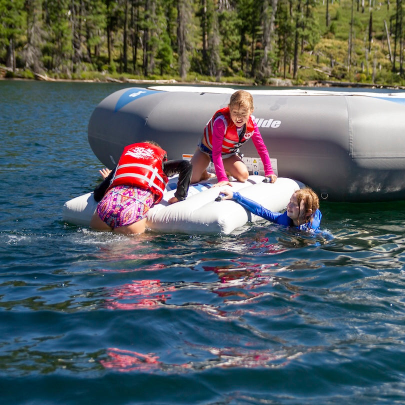 Three kids in life vests use the C-Deck to climb onto an Aquaglide water trampoline.