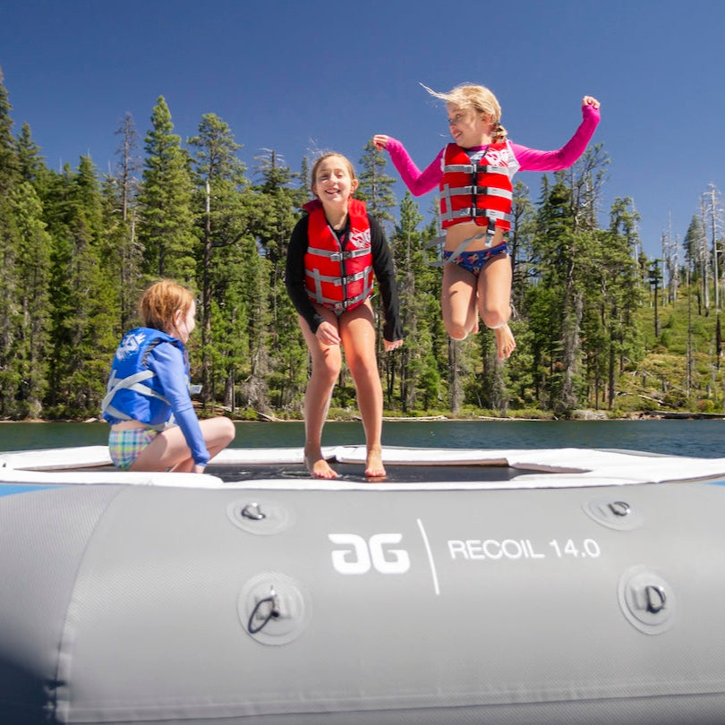 Three kids jumping on the Recoil 14 inflatable water trampoline wearing red and blue life jackets with trees in the background.