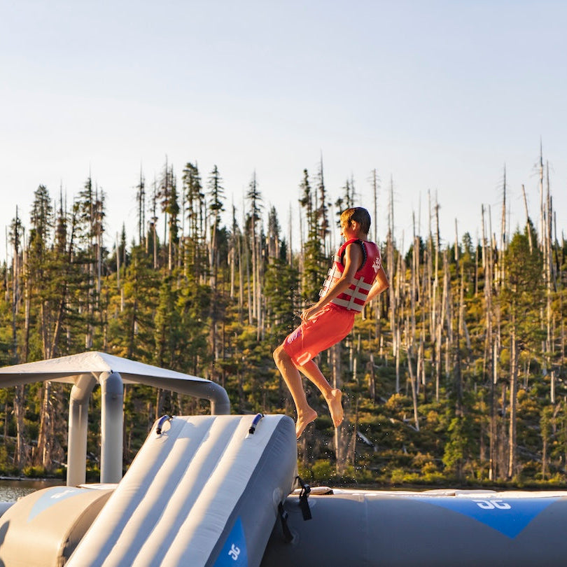 A child in a red life vests plays on a trampoline near the Large Ricochet / Recoil Slide on a lake.