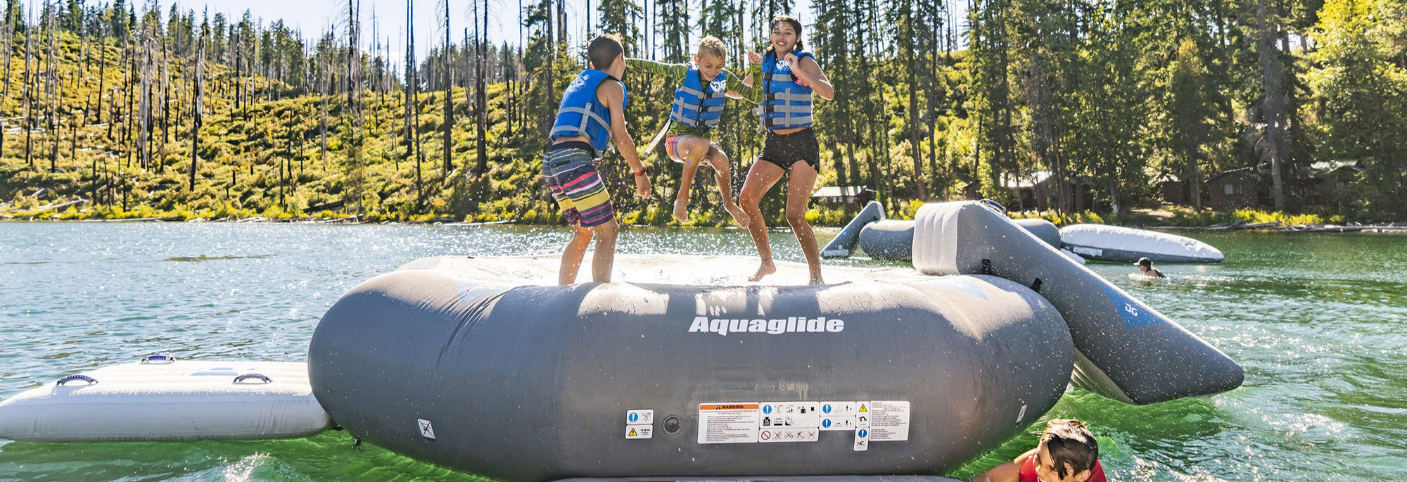 Kids in life vests play on an inflatable water structure in a lake, surrounded by trees.