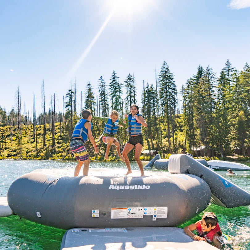 Three kids wearing blue Aquaglide life jackets jumping on the Ricochet 12.0 floating trampoline in the sun with trees in the background.