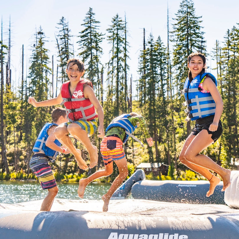 Two kids wearing blue life vests jumping on the Ricochet 12 floating trampoline while two other kids jump into the water wearing blue and red Aquaglide life jackets.