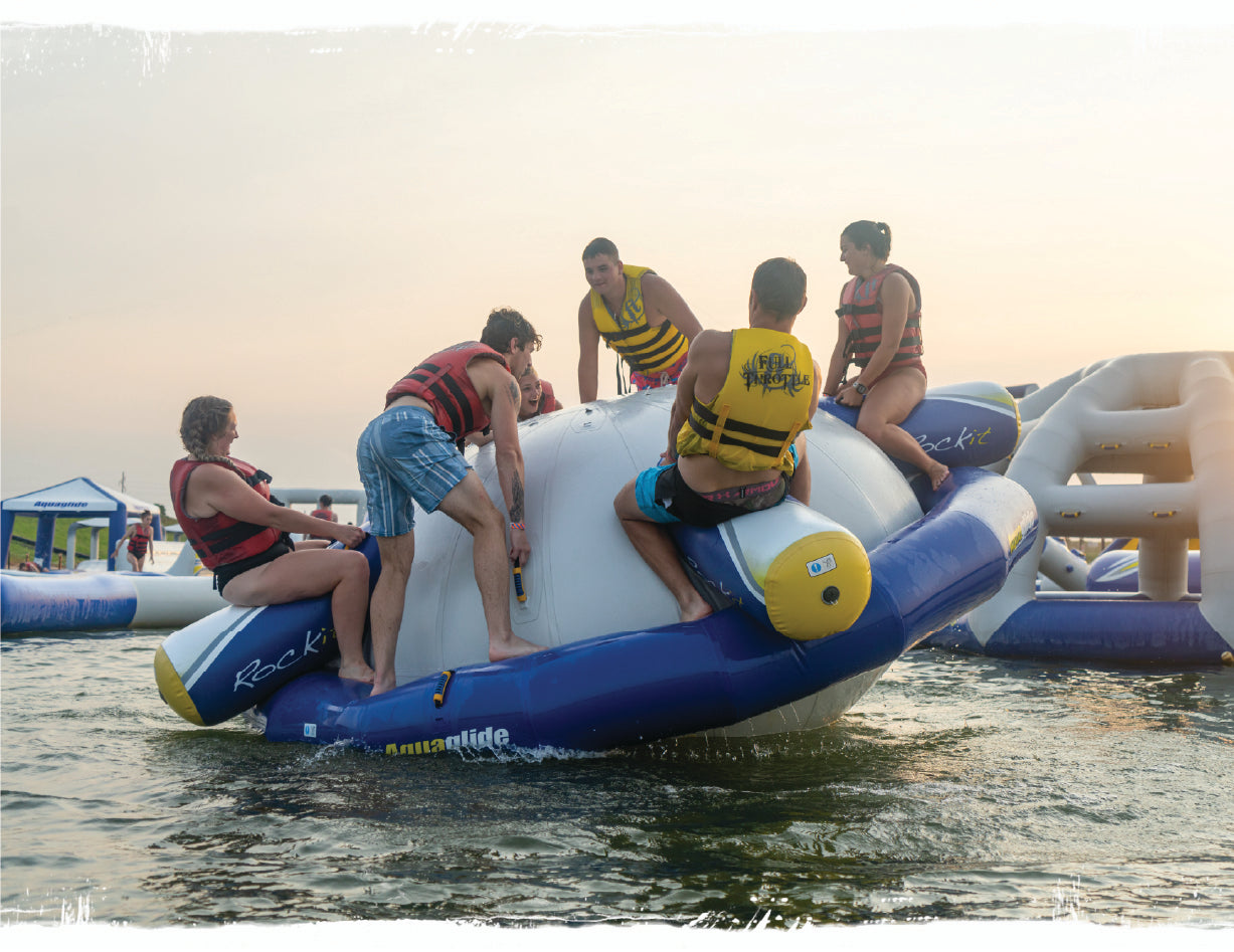 6 swimmers riding the Aquaglide Rock-It trying to knock each other off while wearing red and yellow life jackets