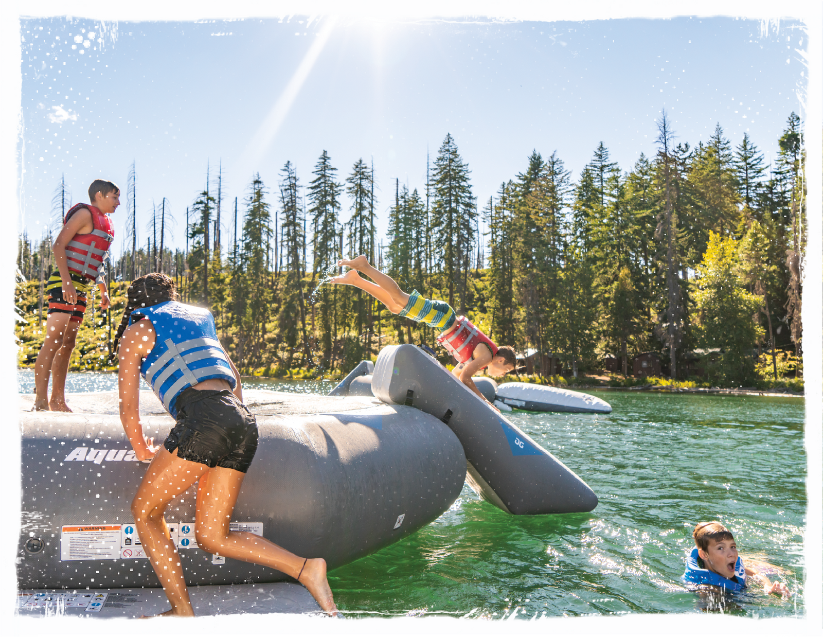 4 kids playing on grey Aquaglide trampoline diving off the slide accessory to swim in the lake.