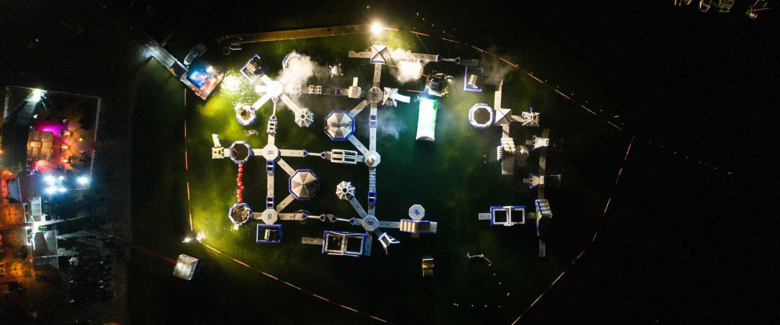 Aerial view of a glowing inflatable obstacle course at night on water.