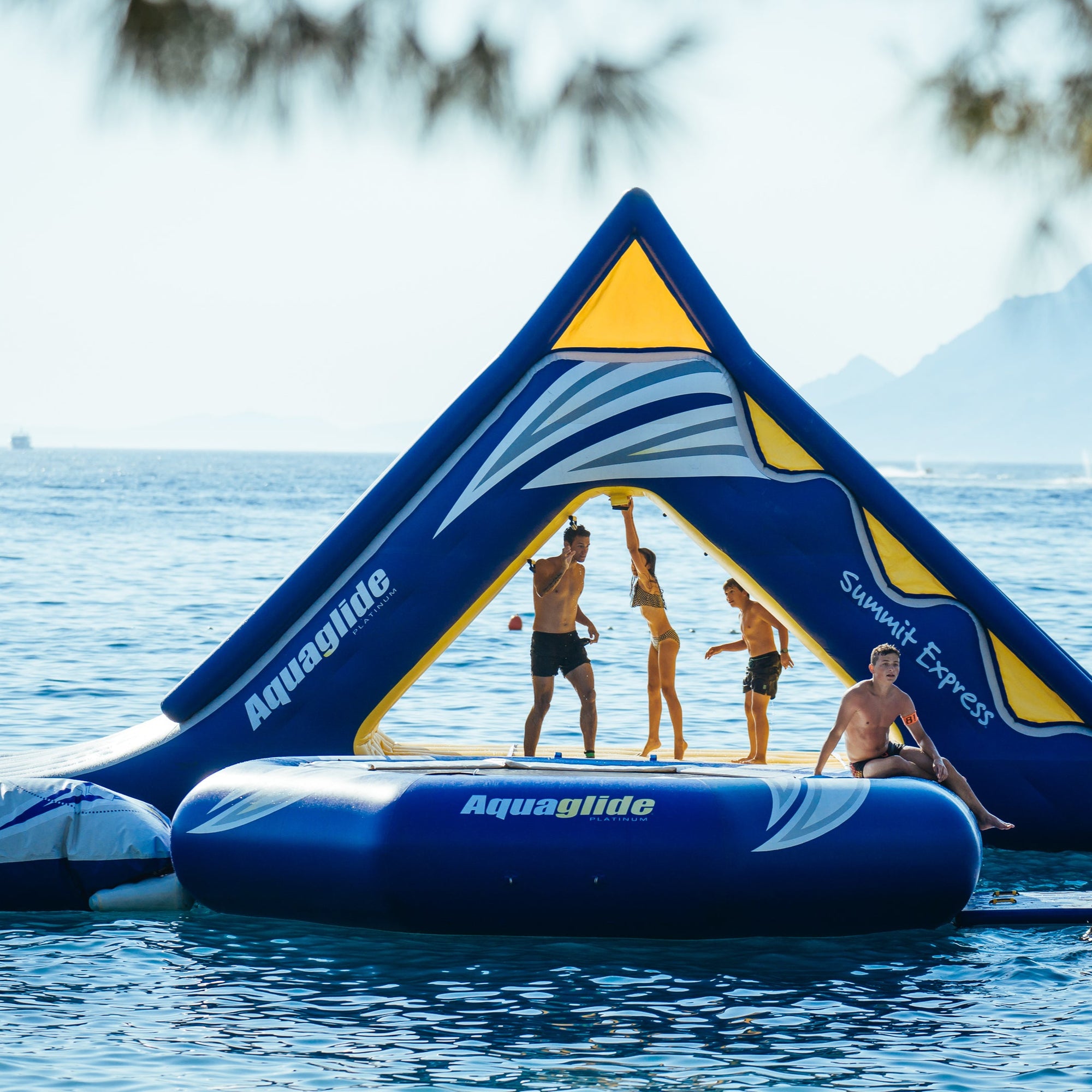 Four people playing on the Aquaglide Summit Express with a blue Aquaglide trampoline on the water. 