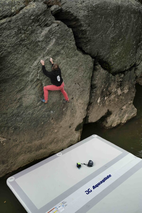 A climber scales a bouldering route over water, with the Landing Pad below to catch their fall