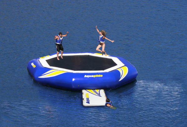 Two adults jumping on a supertramp water trampoline while one boards via the swimstep