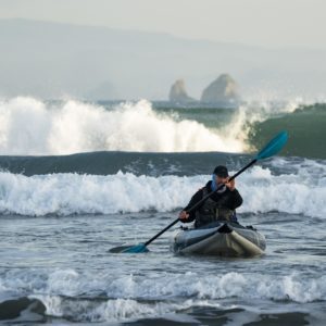 A man paddles a Blackfoot kayak towards shore as large, overhead waves crash in the background
