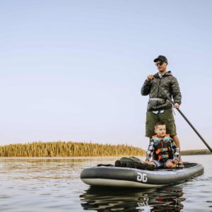 A dad and his son paddle on the Blackfoot Sup. The Dad is standing and paddling while his young son sits and watches