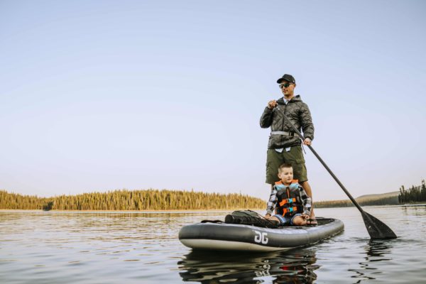 A dad and his son paddle on the Blackfoot Sup. The Dad is standing and paddling while his young son sits and watches