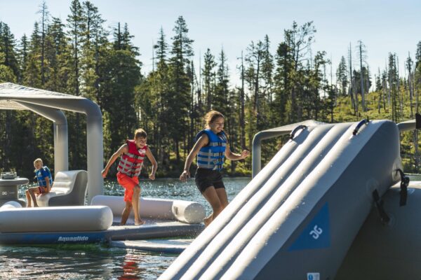 Kids run from an OG lounge to a trampoline in the foreground on a scenic sunny lake