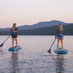 Two woman paddle SUPs away from the camera during sunset, while the pink sky reflects off the water