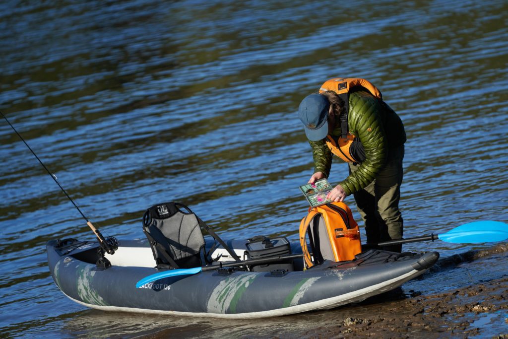A fly fisherman sorting through flies on the beach while standing next to a kayak