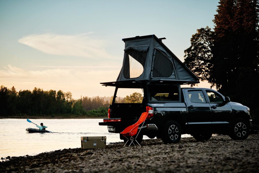 Truck with a canopy camper parked on a gravel bar with a paddler in the background